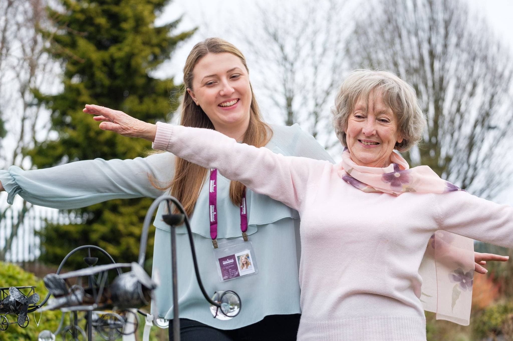 A younger woman and an older woman smiling with outstretched arms outdoors, enjoying their time together. - Home Instead
