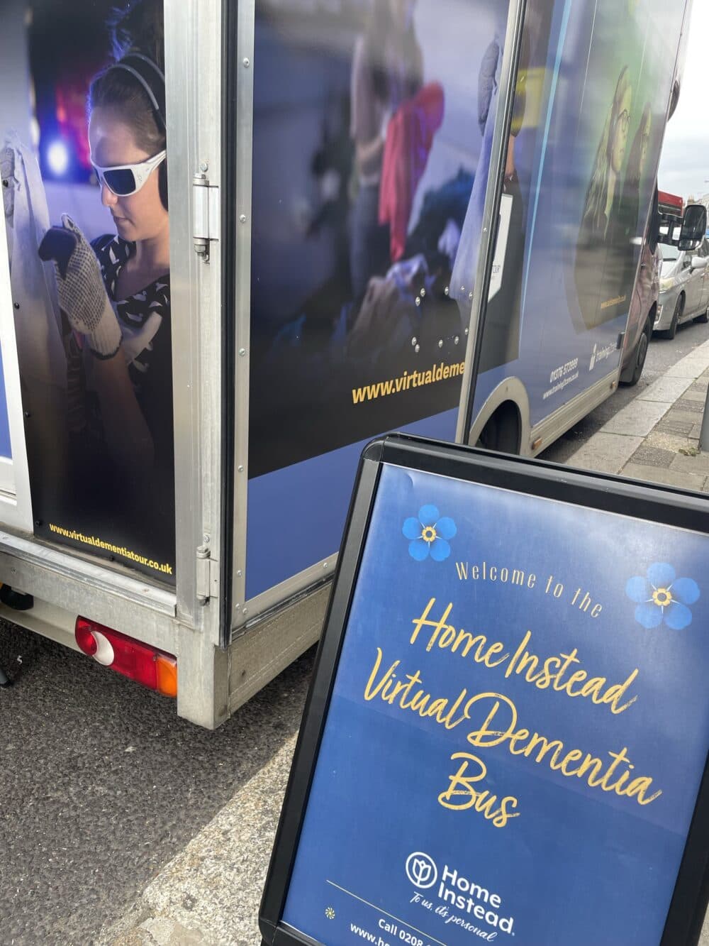 A sign reads "Welcome to the Home Instead Virtual Dementia Bus" in front of the parked bus. - Home Instead