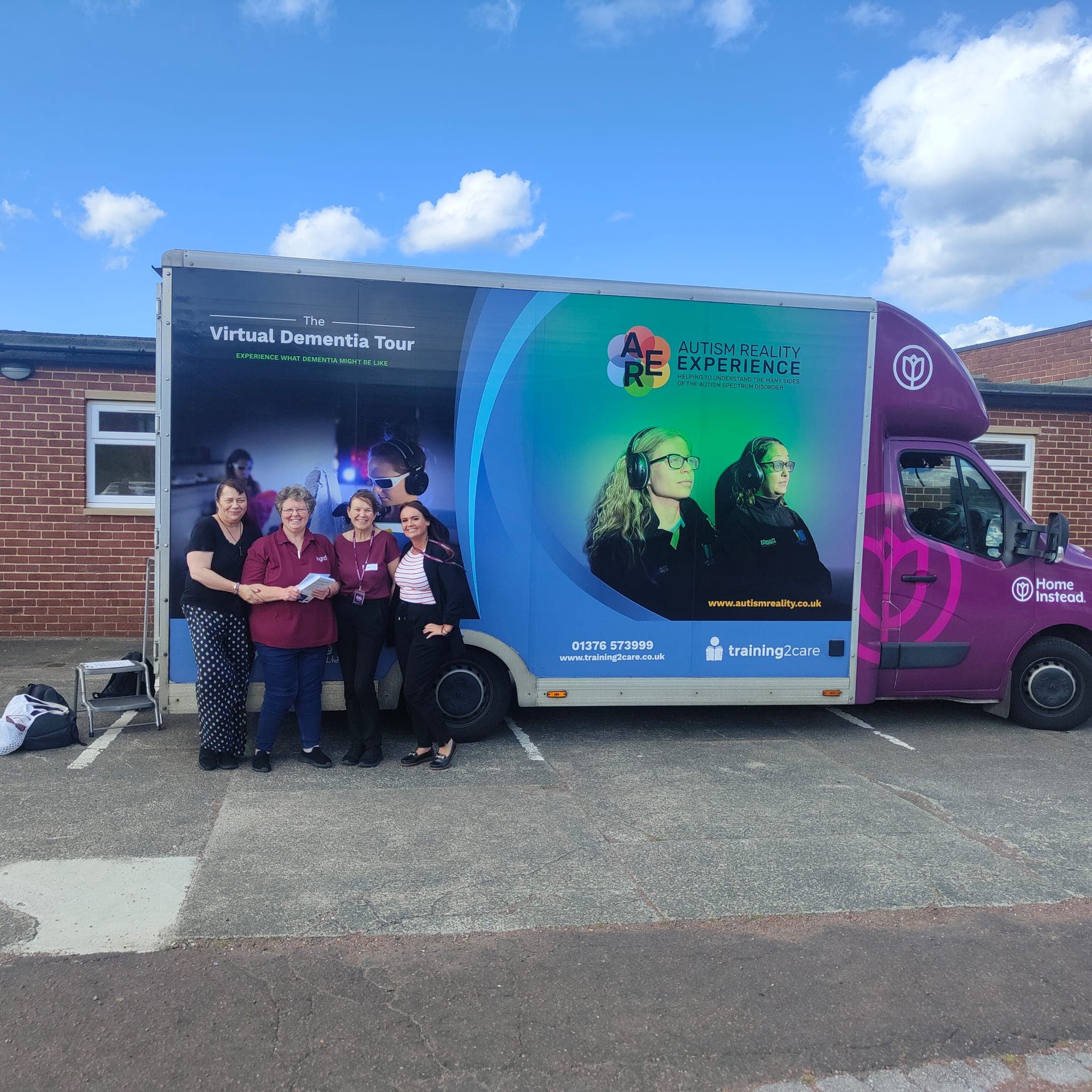 4 people from Home Instead stand outside on a sunny day, in front of the Training 2 Care Dementia Bus