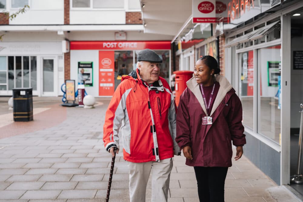 An elderly man with a cane walks with a caretaker outside a post office. - Home Instead