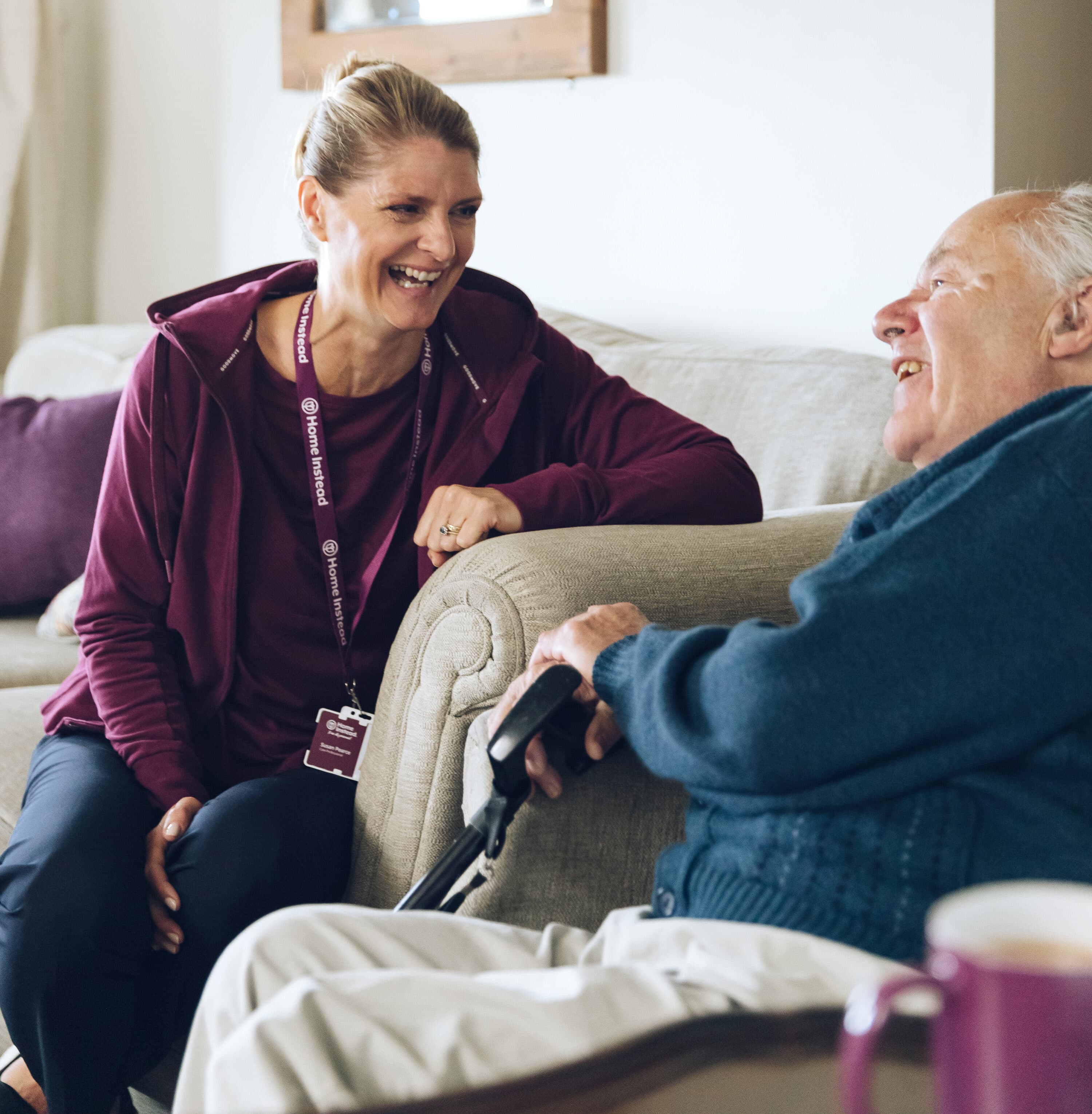 A woman in a purple shirt chats and laughs with an elderly man sitting on a couch, holding a walking cane. - Home Instead