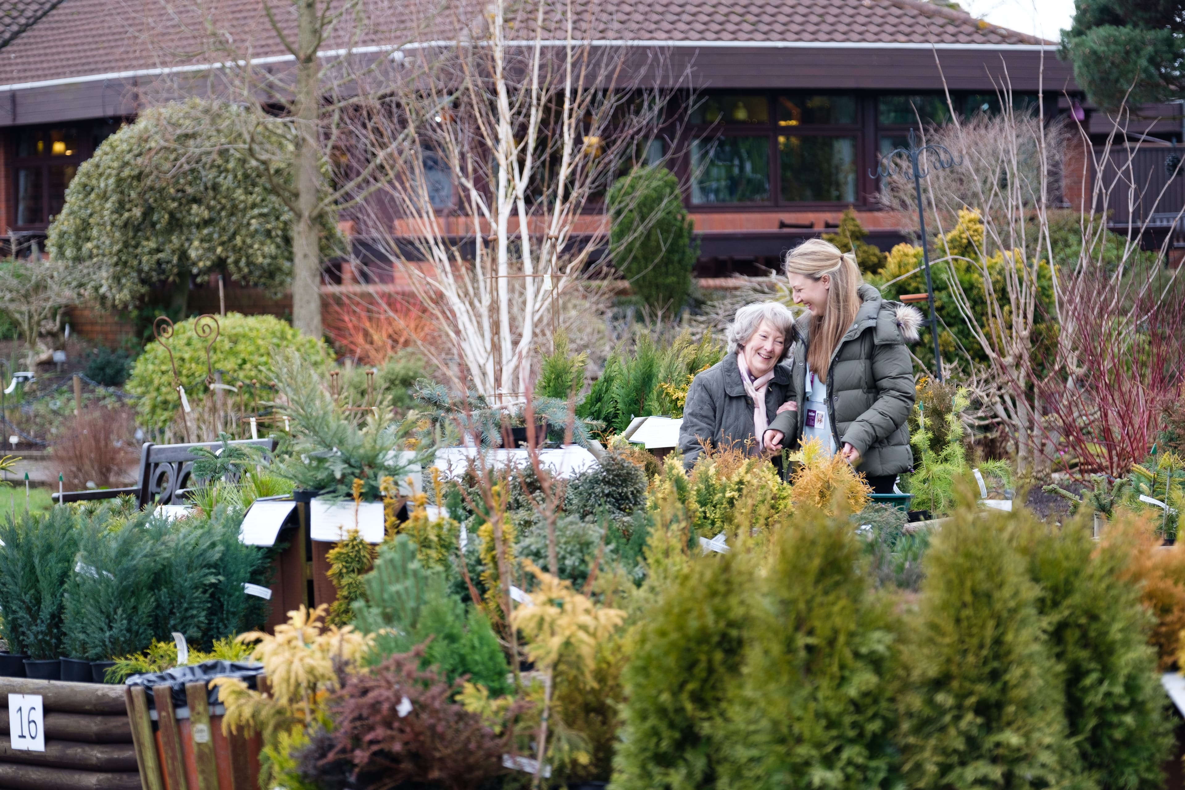 Two women, one elderly and one younger, enjoy looking at plants in a garden center on an overcast day. - Home Instead