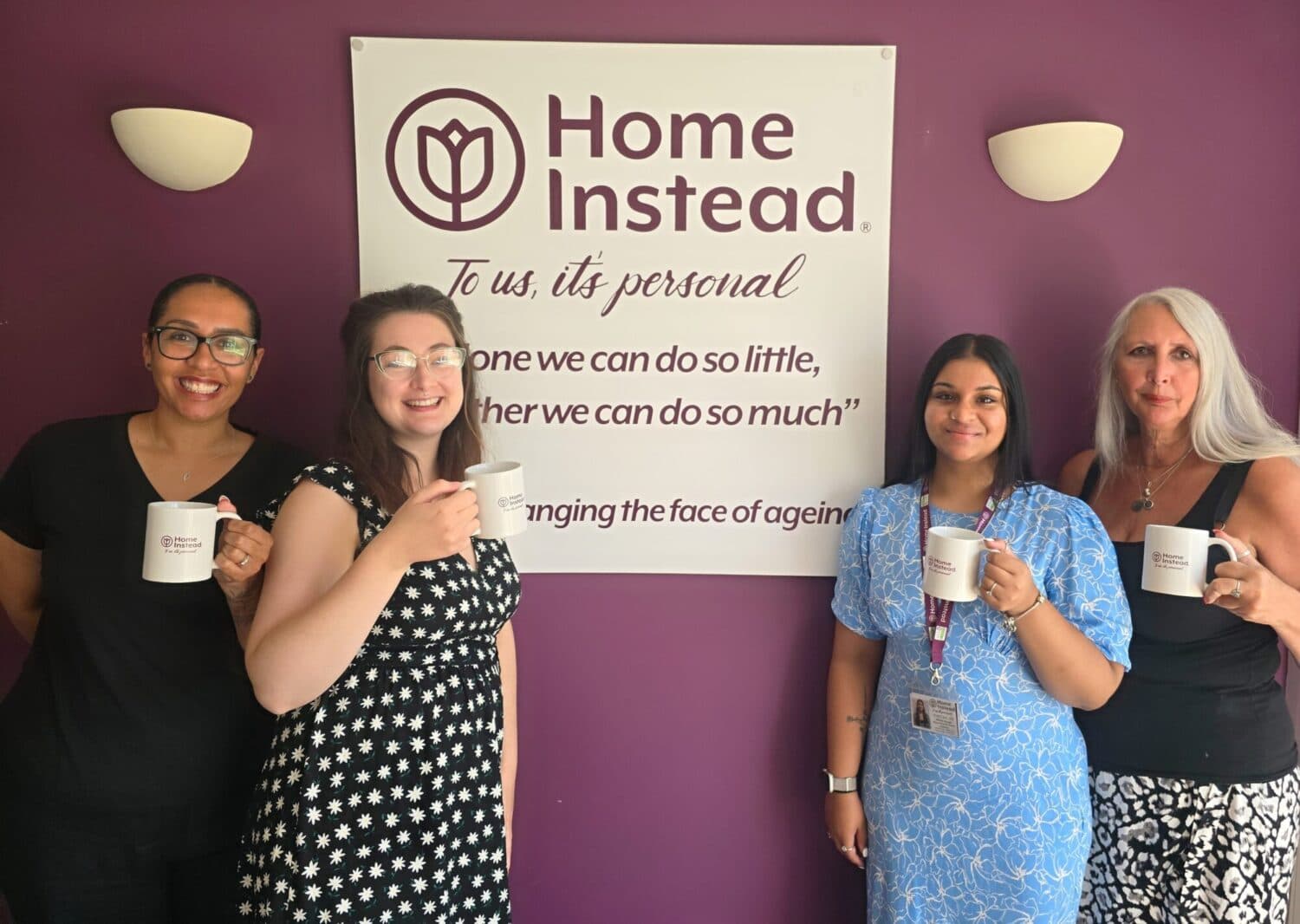 Four women holding mugs, standing in front of a "Home Instead" sign on a purple wall. - Home Instead