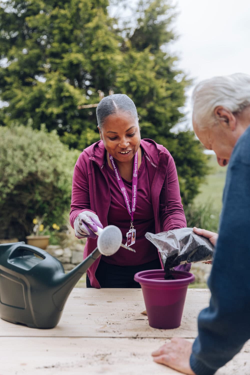 Two people gardening together; one is pouring soil into a purple pot on a wooden table. - Home Instead