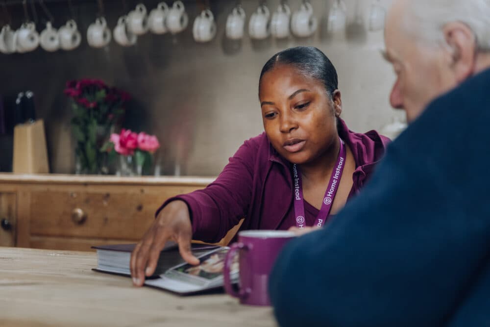 A woman shows a book to an older man at a kitchen table with a purple mug and flowers in the background. - Home Instead