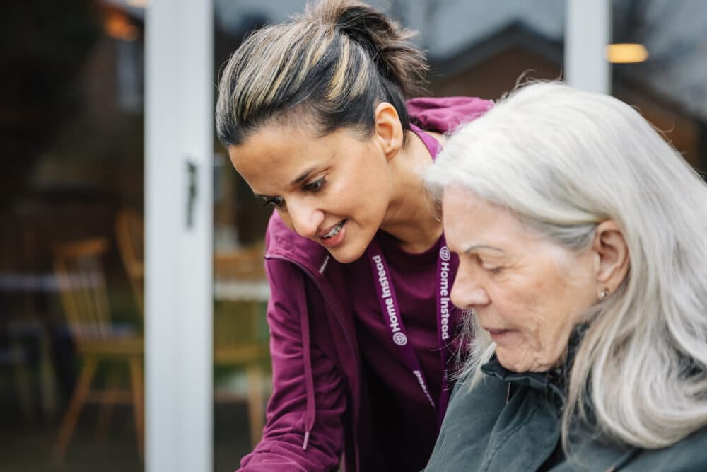 A caregiver assists an elderly woman outdoors, both smiling. - Home Instead