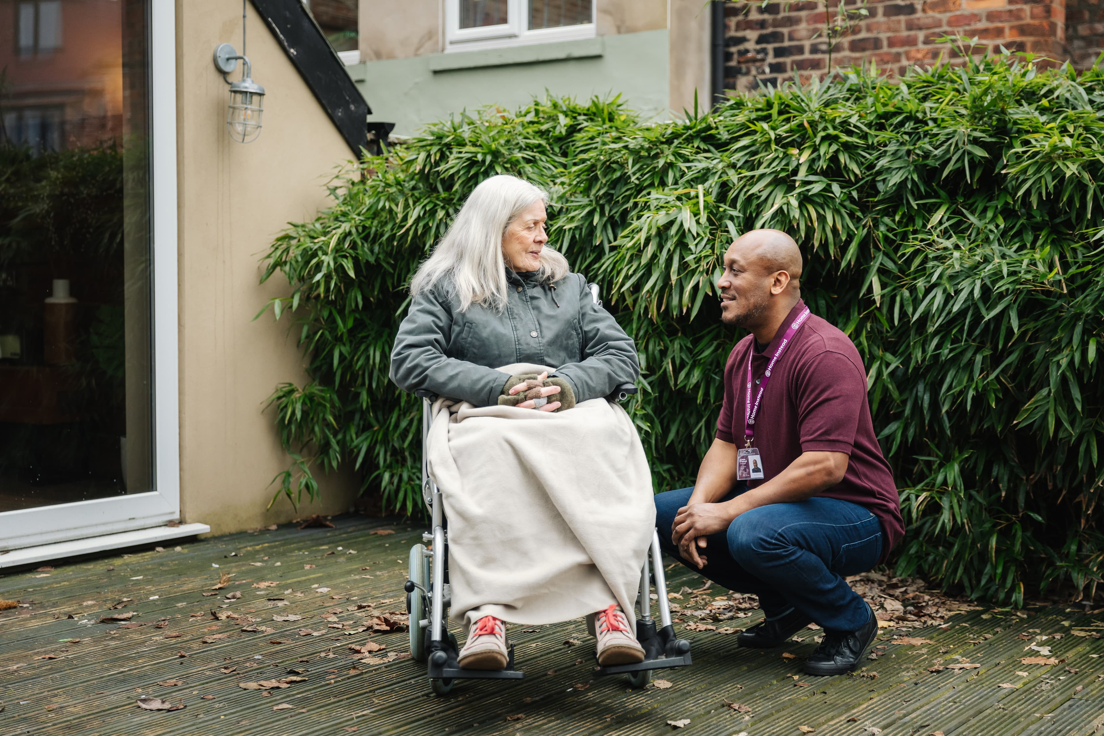 A caregiver kneels and smiles, talking to a woman in a wheelchair outdoors. - Home Instead