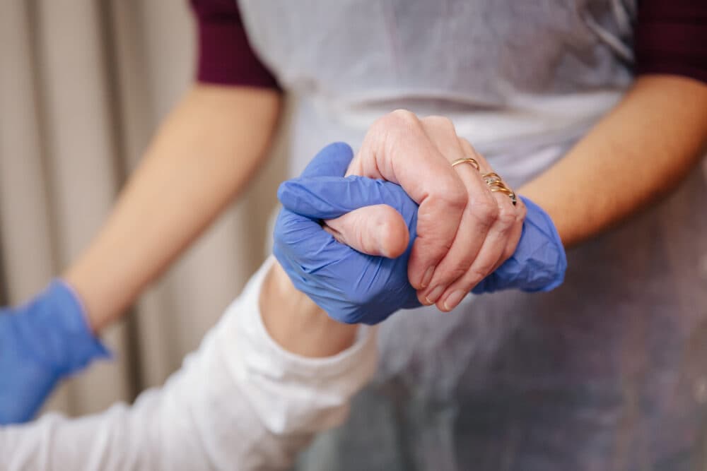 Two people clasp hands, one wearing blue gloves, symbolizing care and support. - Home Instead