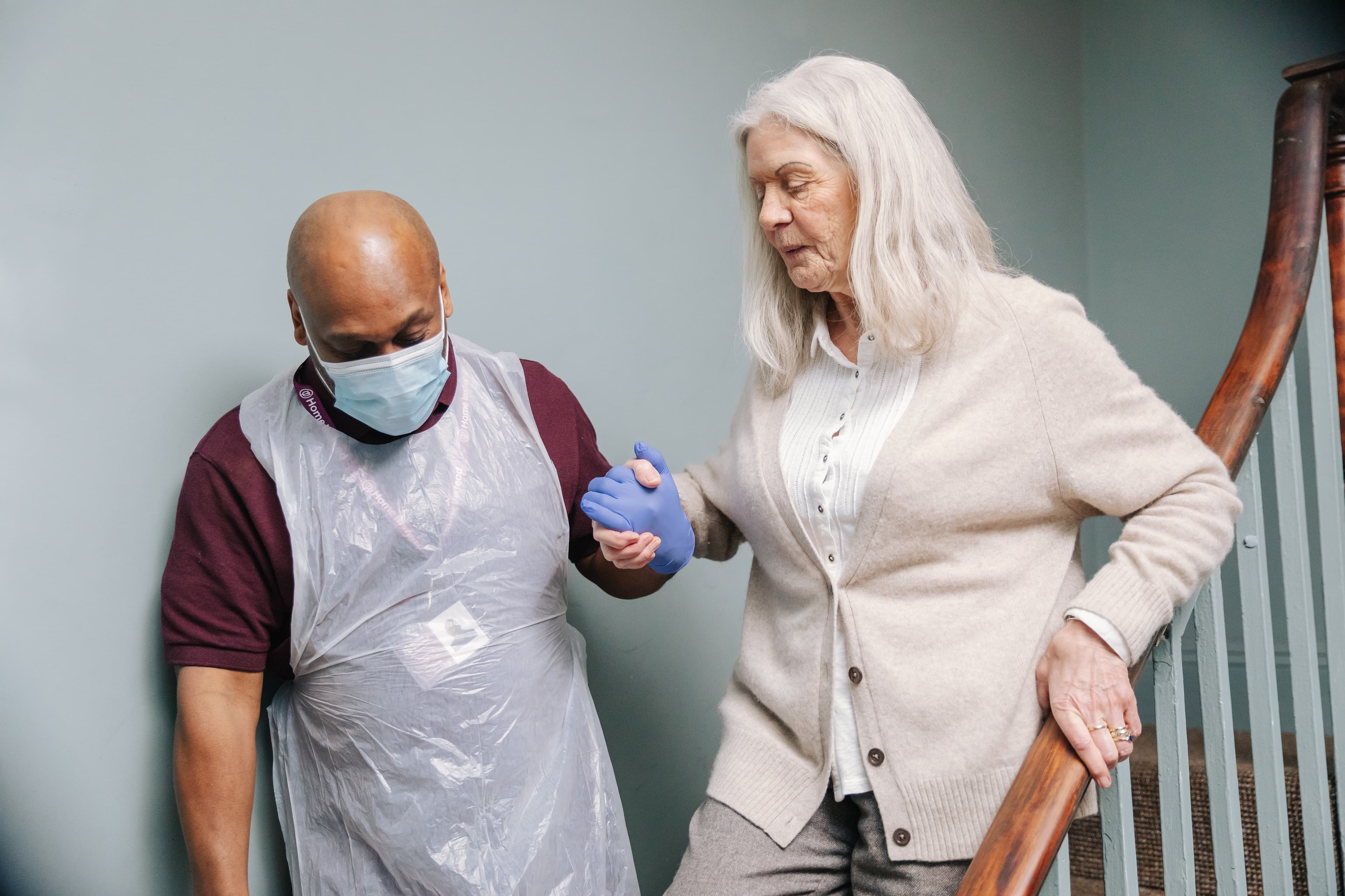 A caregiver wearing a mask assists an elderly woman down the stairs. - Home Instead