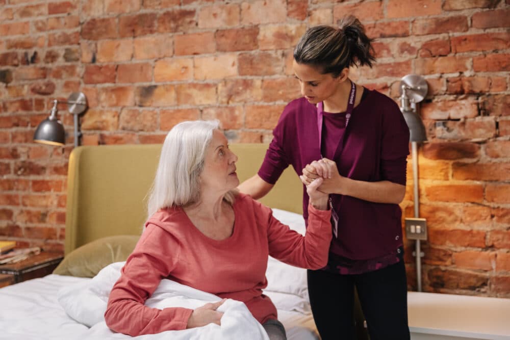 Caregiver helping an elderly woman out of bed, with a brick wall background. - Home Instead