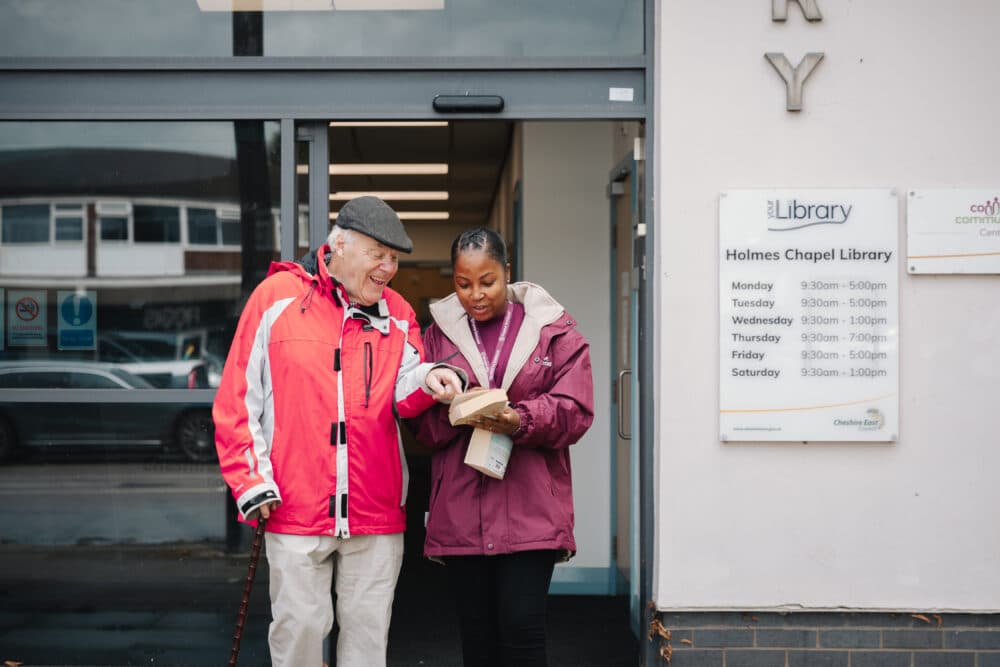 Two people in jackets stand outside a library, looking at a book together. - Home Instead