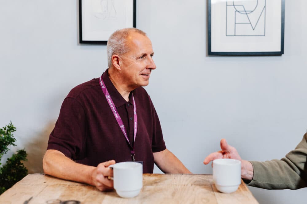 Man in a maroon shirt holding a mug, seated at a wooden table talking to another person, with framed art on the wall. - Home Instead