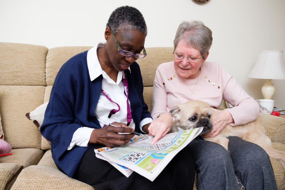 A woman with glasses helps an older woman with gray hair complete a crossword puzzle while a dog sits on their laps. - Home Instead