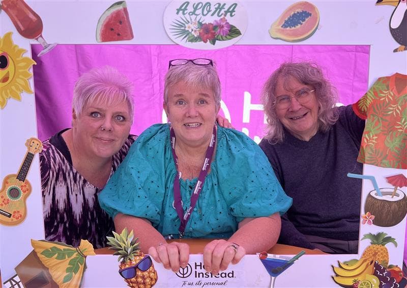 Three smiling adults pose together behind a colorful tropical-themed photo frame with "Aloha" at the top. - Home Instead