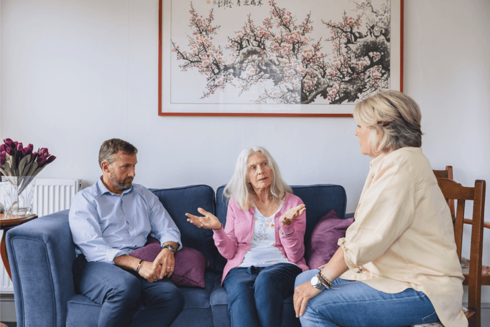 Three people sitting in a living room, engaged in a serious conversation, with floral artwork on the wall behind them. - Home Instead