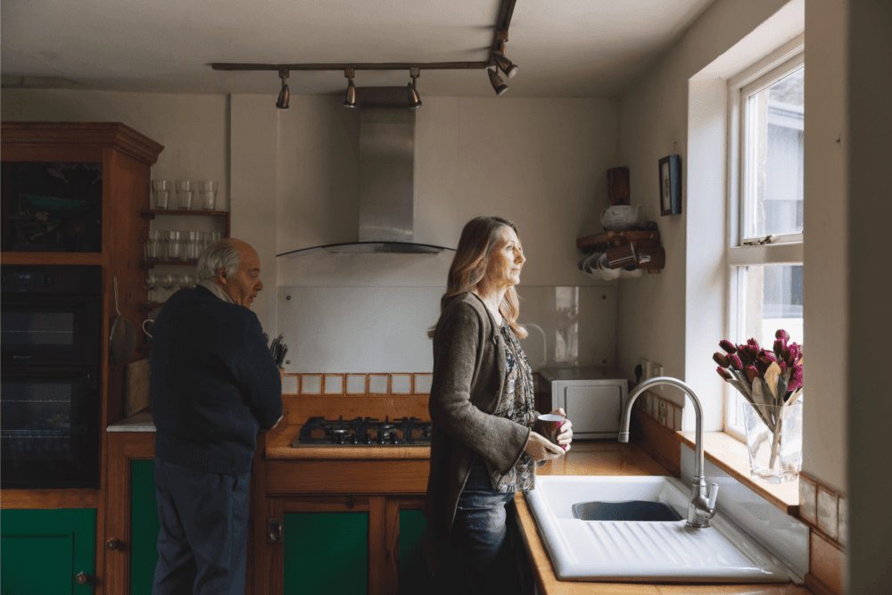 A woman holding a mug looks out of a kitchen window while a man stands at the counter in the background. - Home Instead