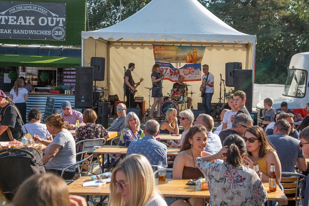 Outdoor music event with people seated at tables, a band performing on stage, and a food stand in the background. - Home Instead