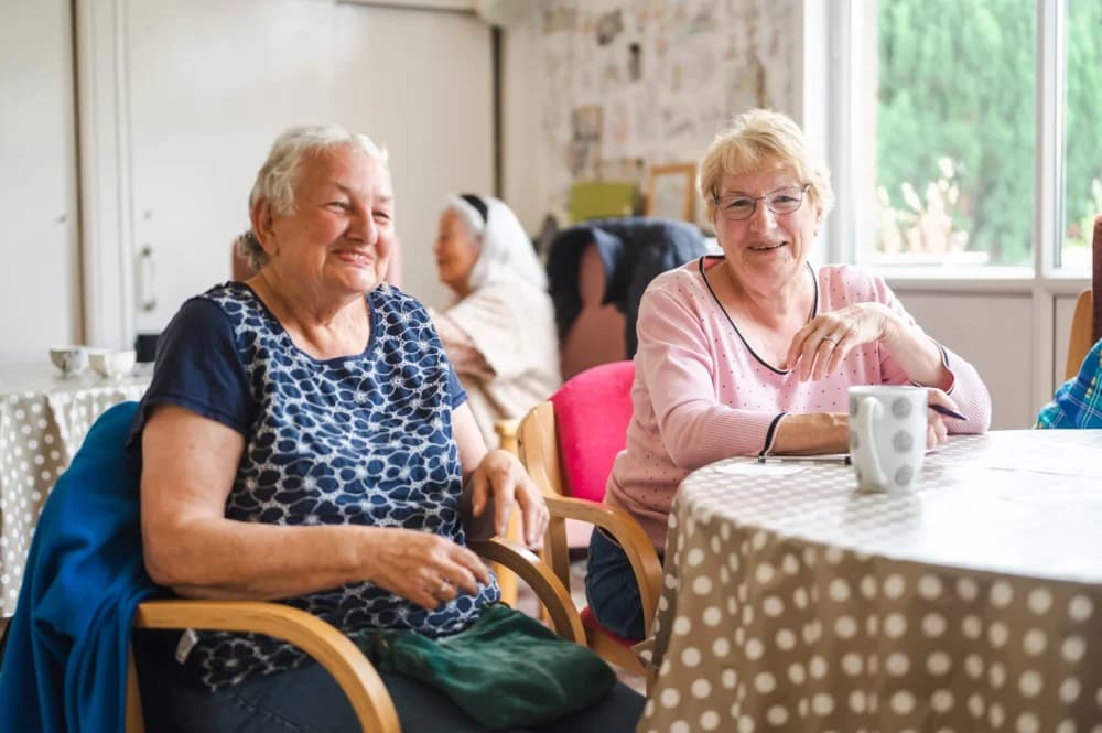 Two elderly women sitting at a table with a polka-dot tablecloth, smiling and talking in a bright, cozy room. - Home Instead