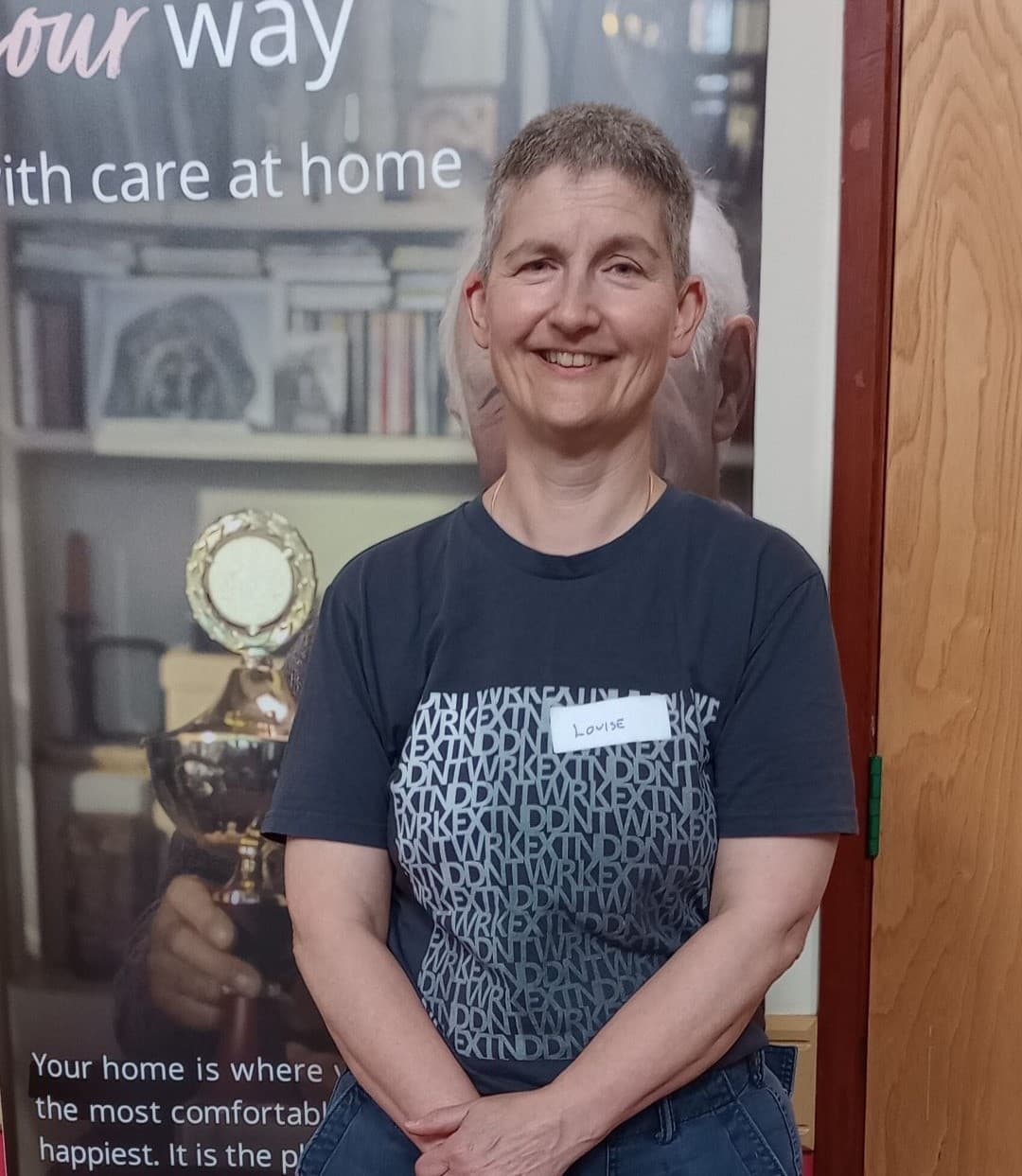 A person with short hair smiles in front of a display. They are wearing a nametag that says "Louise. - Home Instead