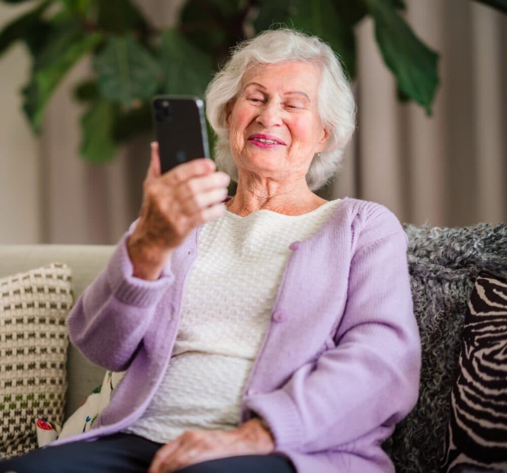 Elderly woman with white hair and a lavender cardigan smiling while looking at a smartphone. - Home Instead