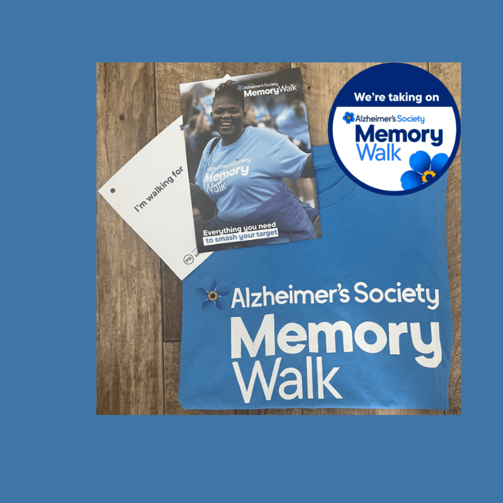 Alzheimer's Society Memory Walk t-shirt, pamphlet, and badge on a wooden surface. - Home Instead
