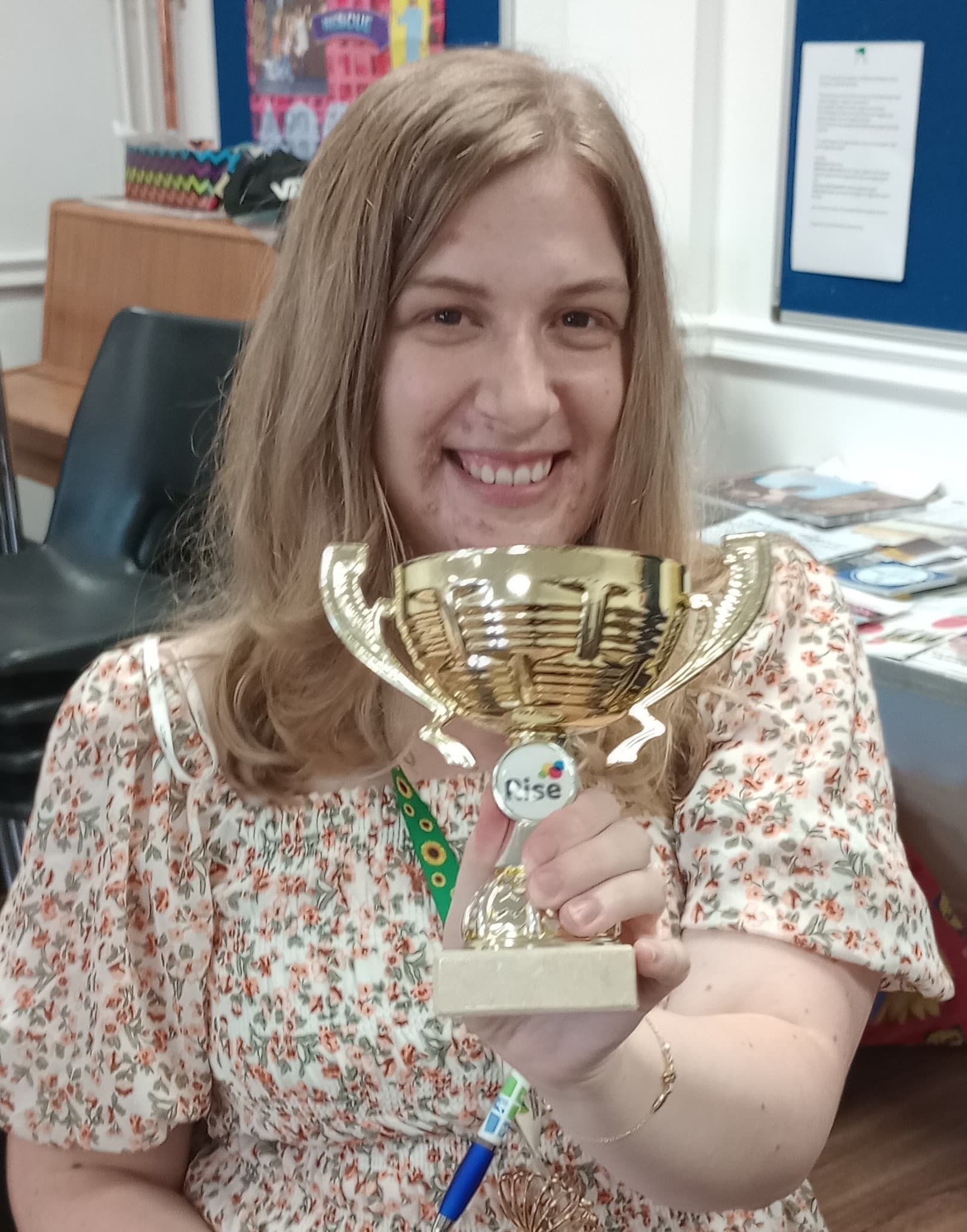 A smiling woman holding a gold trophy in a room with chairs and a bulletin board in the background. - Home Instead