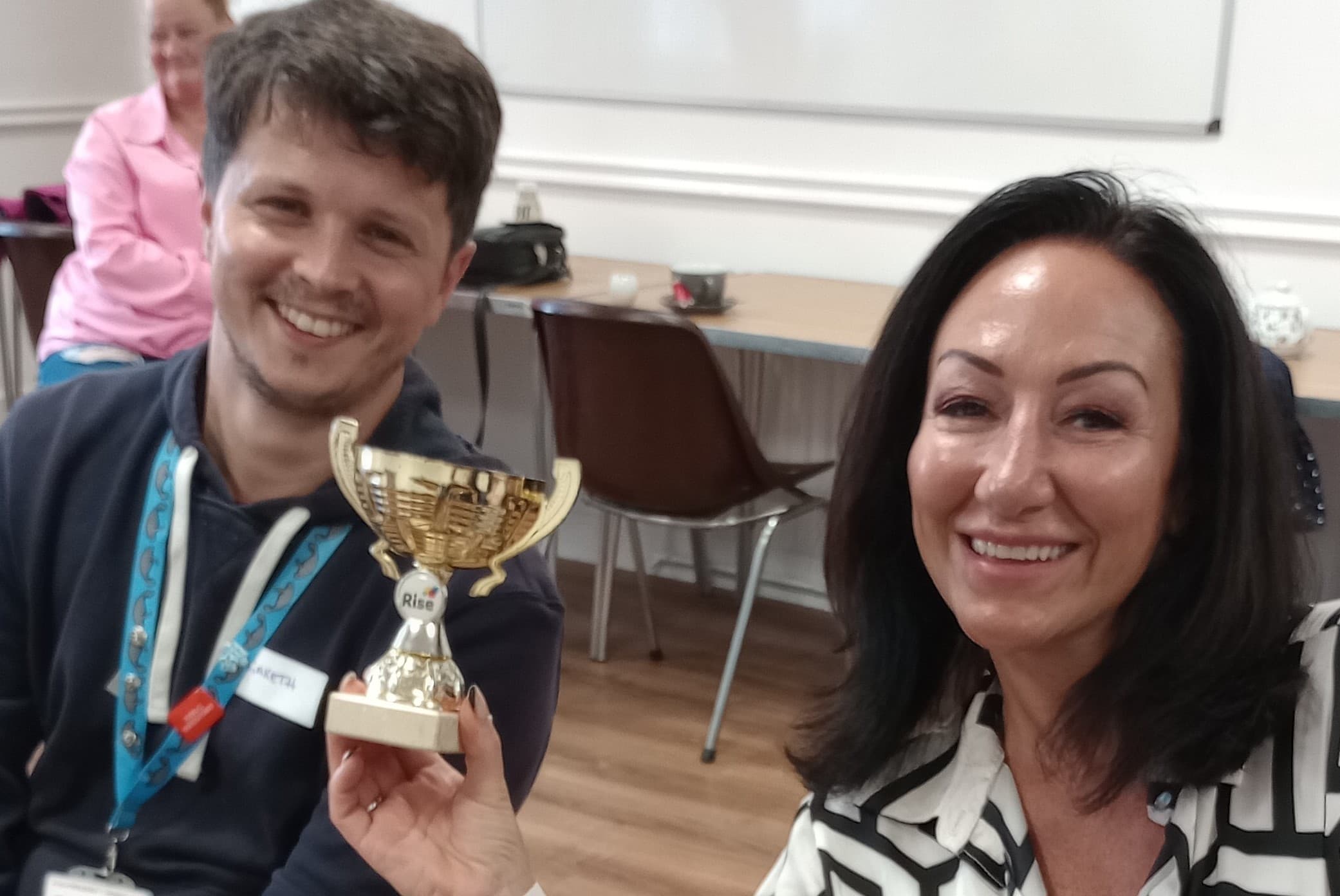 Two smiling people, one holding a gold trophy with "1st" on it, seated in a room with a whiteboard in the background. - Home Instead