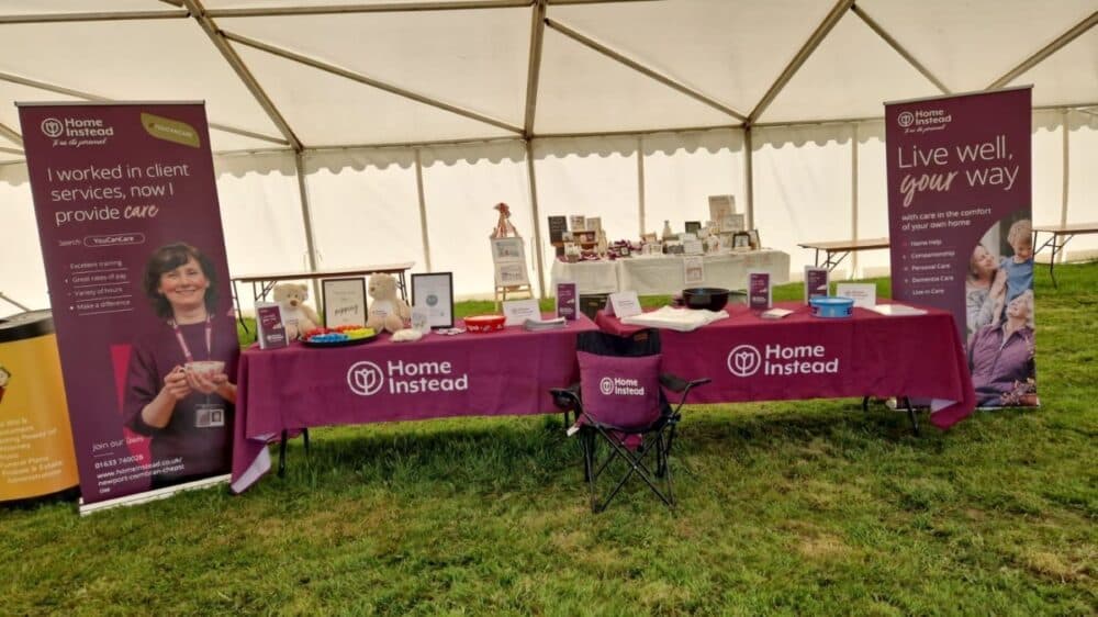 A Home Instead stall under a tent, showcasing banners, informational materials, and merchandise on tables. - Home Instead