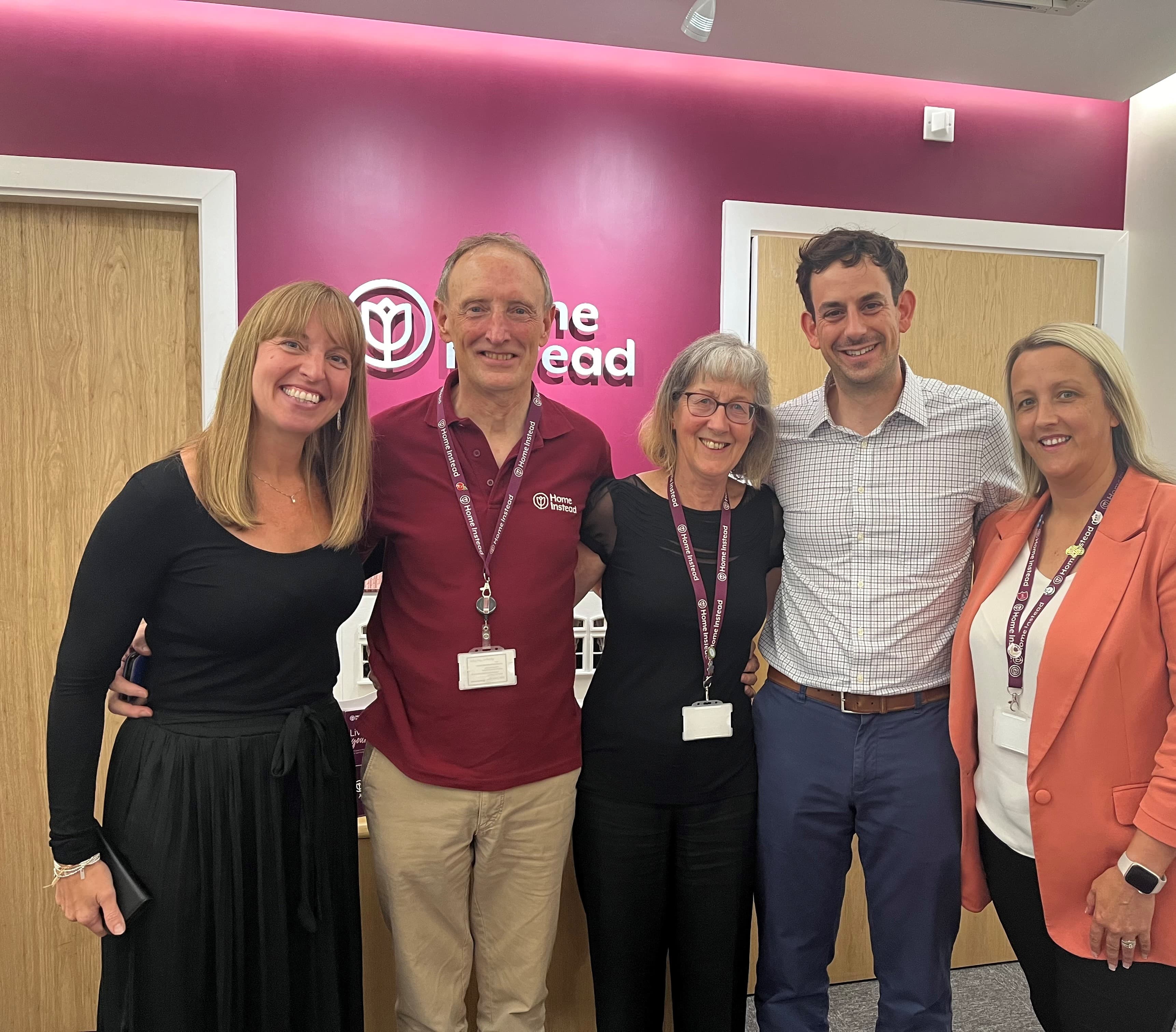 Five people smiling, standing in a group in front of a pink wall with a logo and two doors. - Home Instead