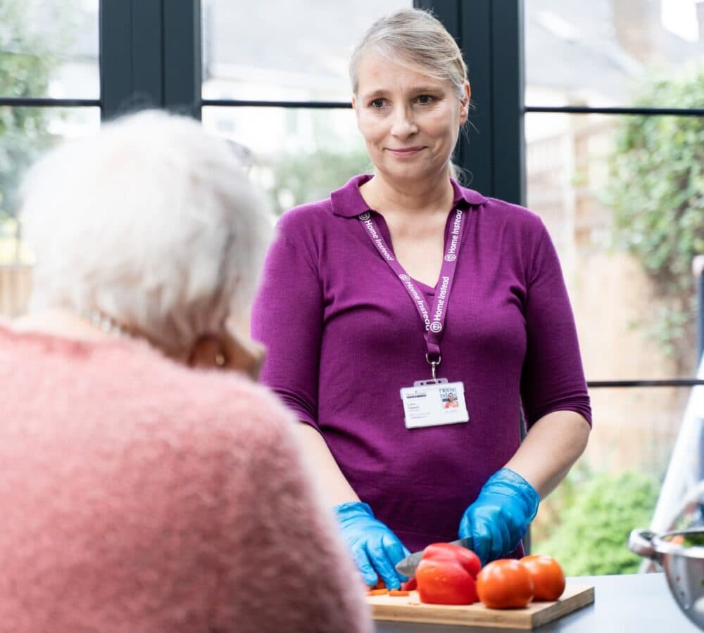 Woman in purple shirt, wearing gloves, cuts tomatoes while talking to an elderly person in a pink sweater. - Home Instead