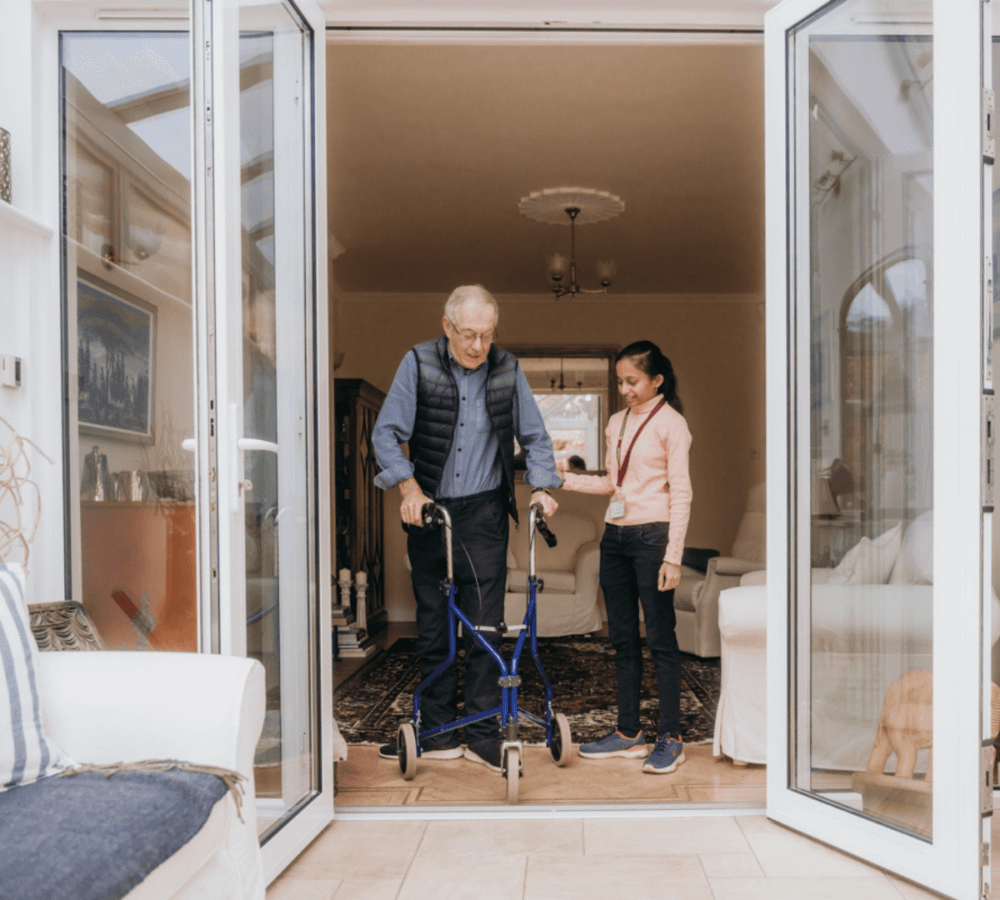 An elderly man using a walker is assisted by a young woman as they move through a doorway at home. - Home Instead