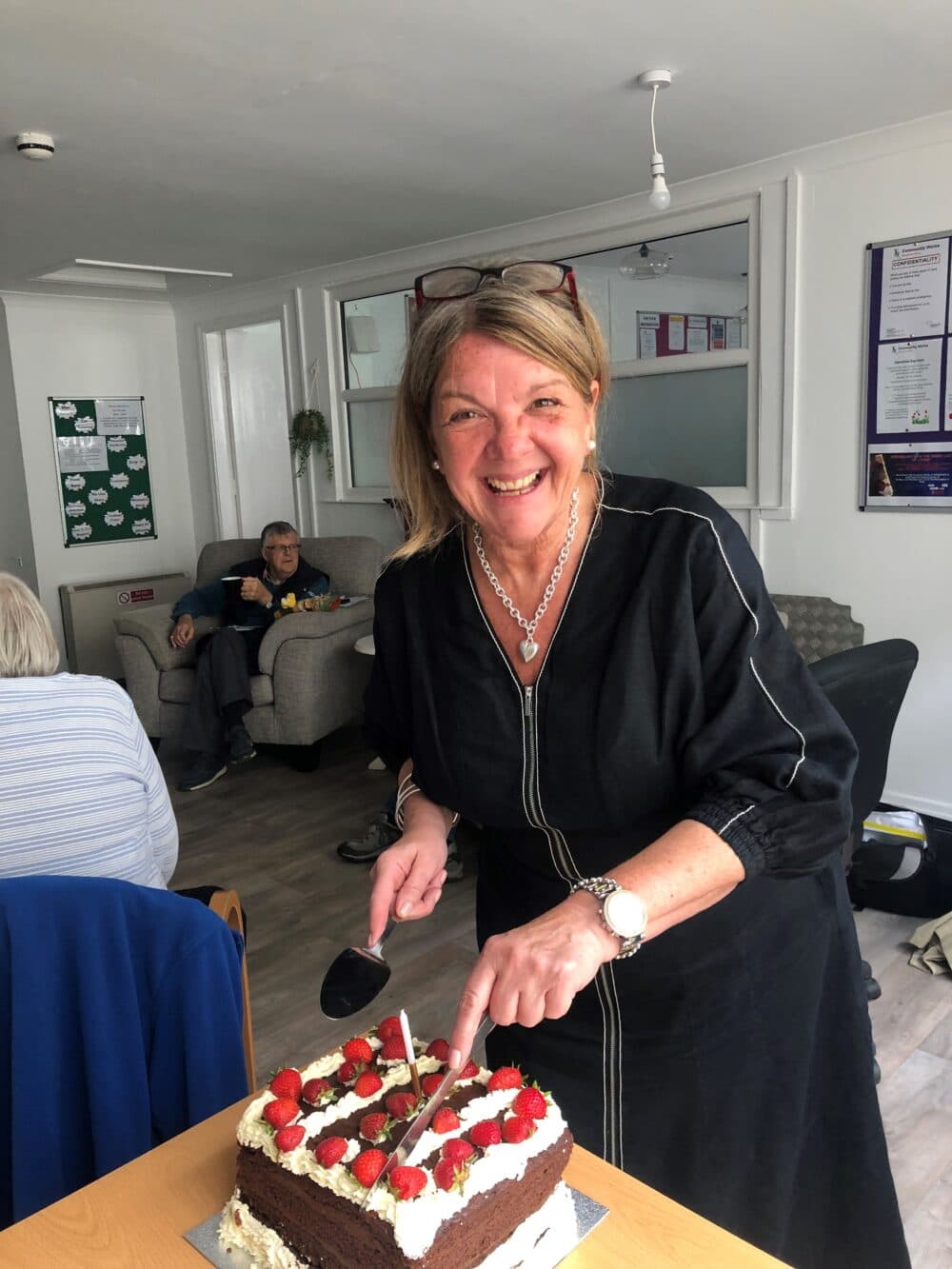 A woman in a black dress smiles while cutting a cake with strawberries and cream in a room with people in the background. - Home Instead