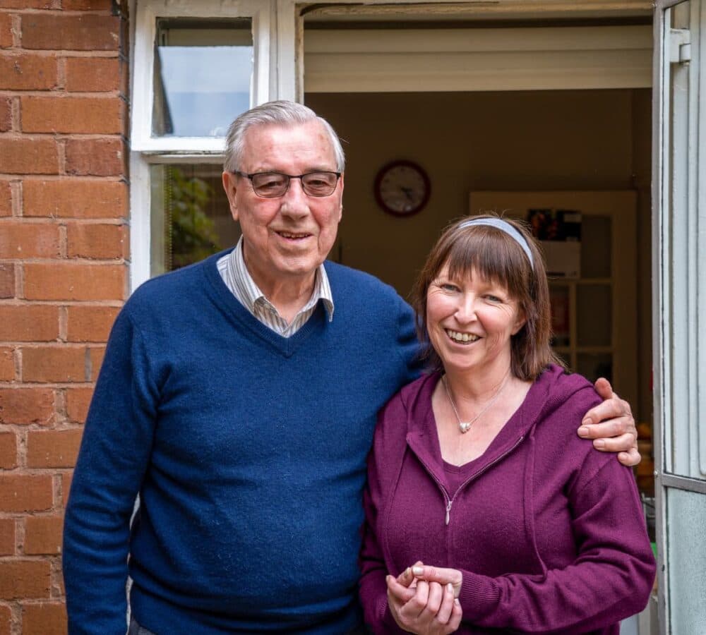 An elderly man and a middle-aged woman smiling, standing in front of an open door next to a brick wall. - Home Instead