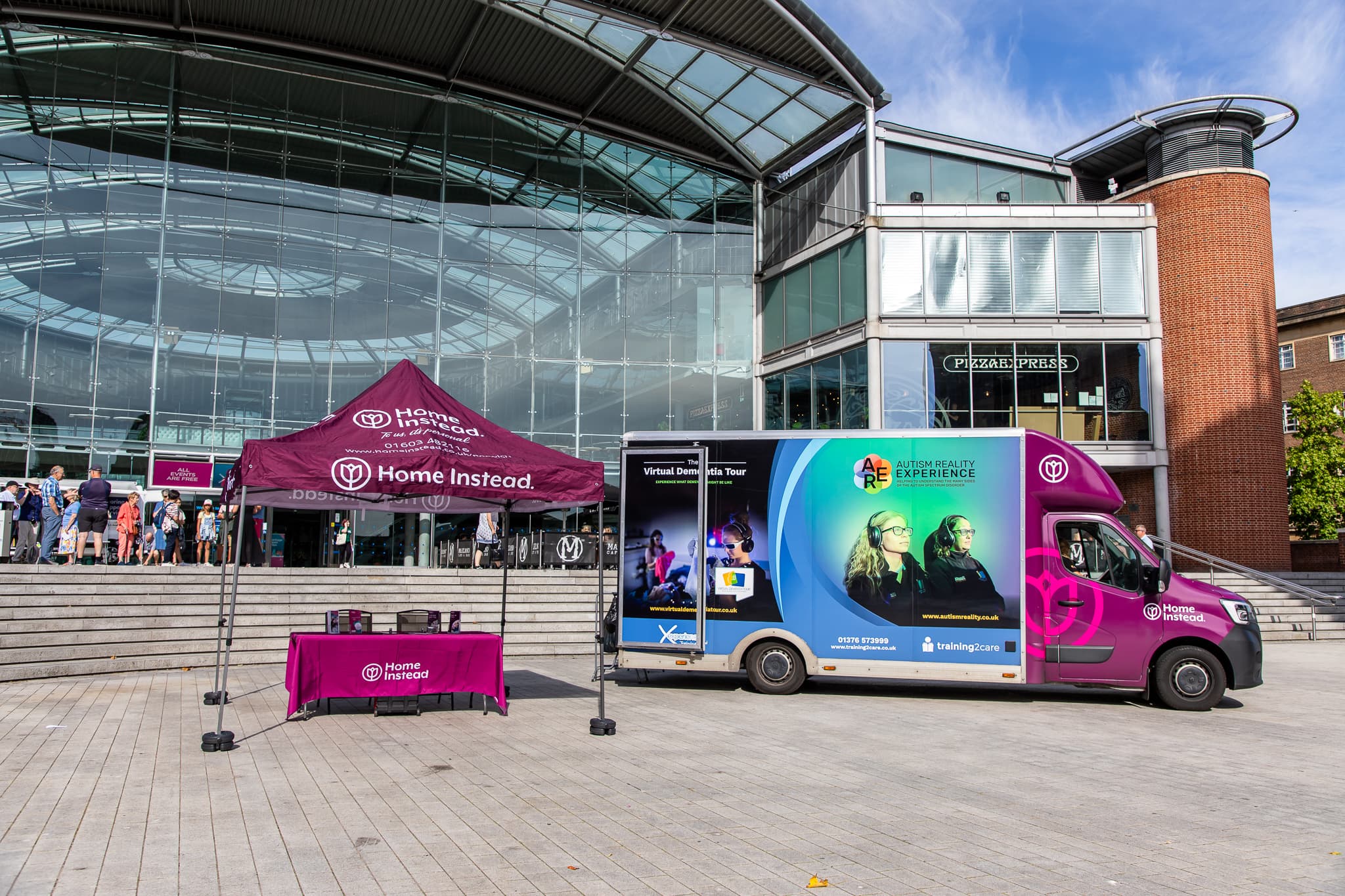 Mobile exhibit and tent outside a modern glass building with people ascending steps. - Home Instead