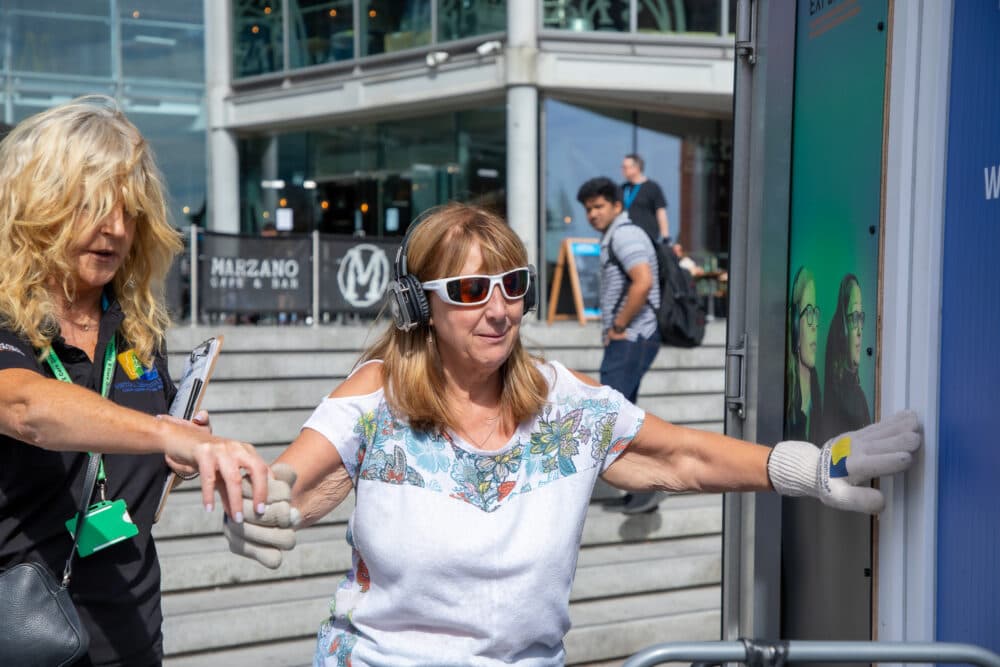 A woman with headphones and white sunglasses balances with assistance outside a building. - Home Instead