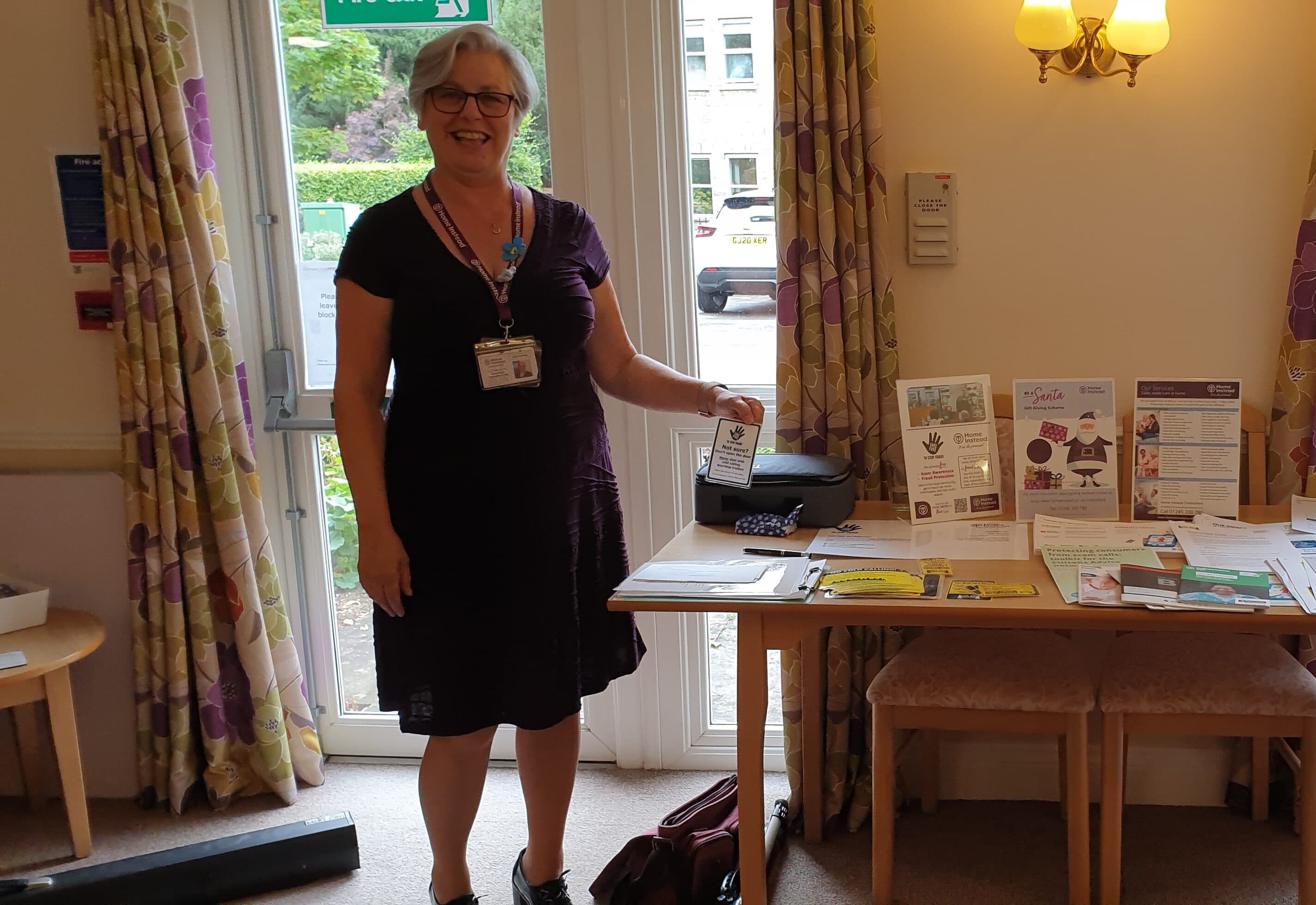 A woman standing in front of a table with brochures and posters in a well-lit room. - Home Instead