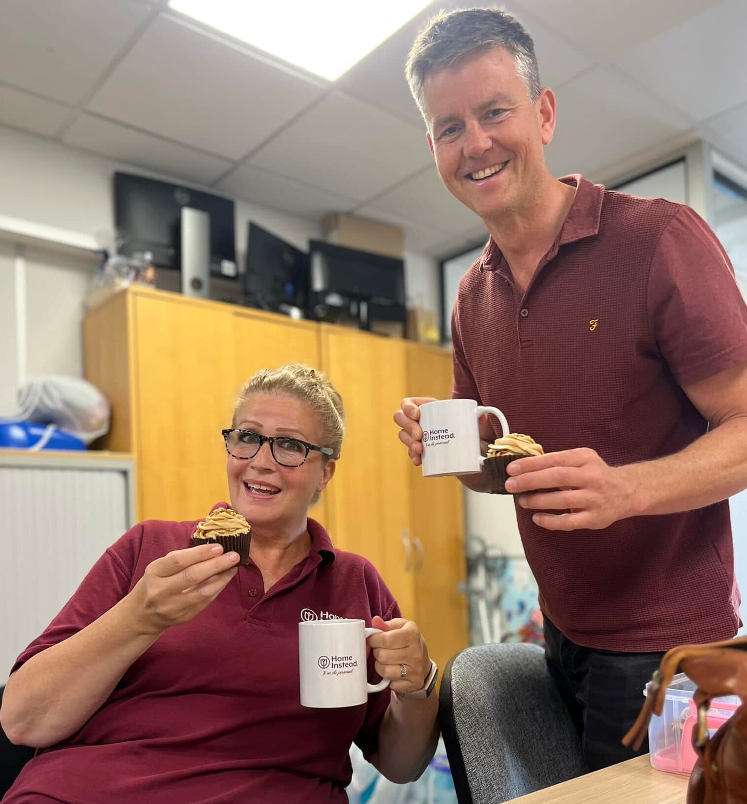 Two people smiling, holding mugs and cupcakes, sitting in an office with a cabinet and computers in the background. - Home Instead