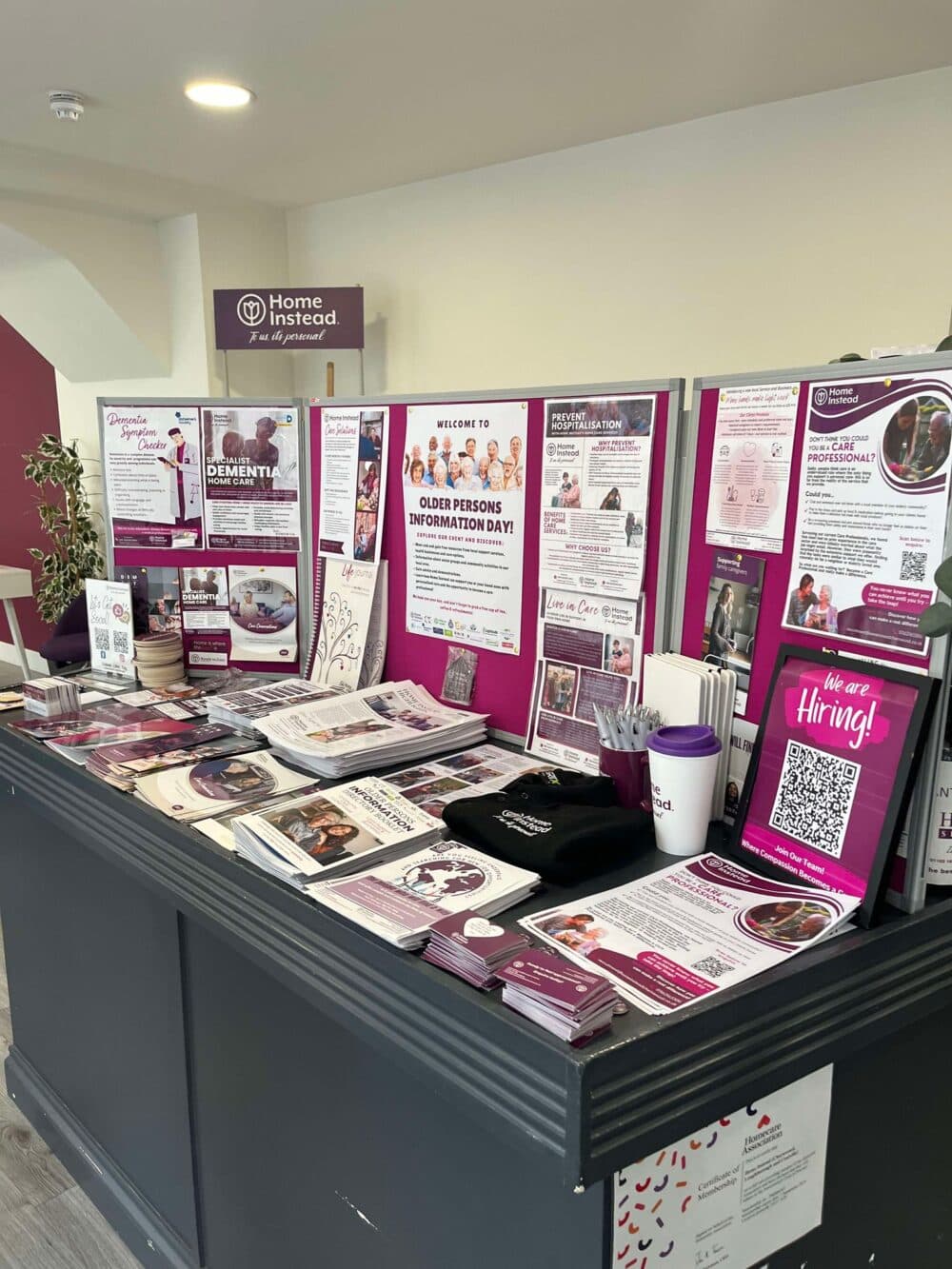 A display table with flyers, brochures, and a "We're Hiring" sign at an Older Persons Information Day event. - Home Instead