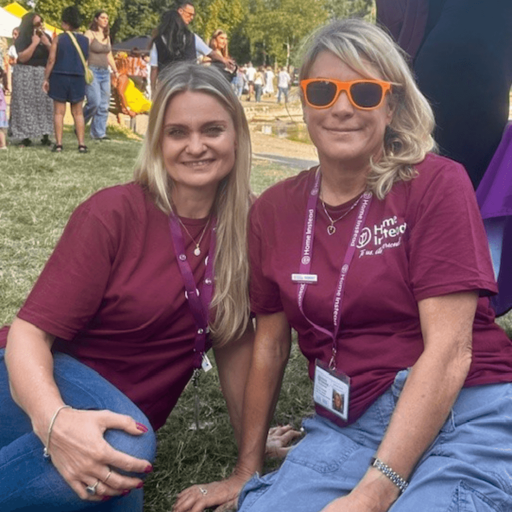 Two women in maroon shirts and lanyards smiling outdoors at a park event. - Home Instead