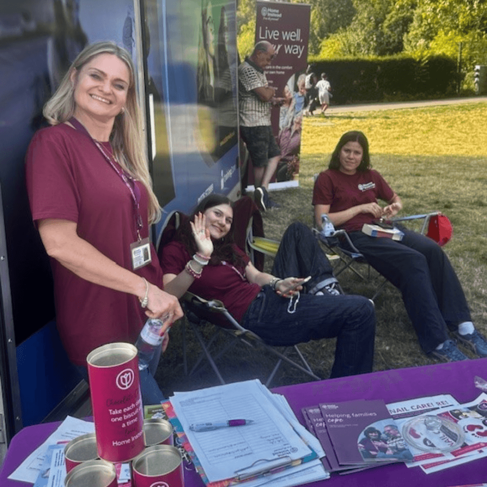 Three people in maroon shirts at an outdoor information booth with brochures and donation cans on the table. - Home Instead