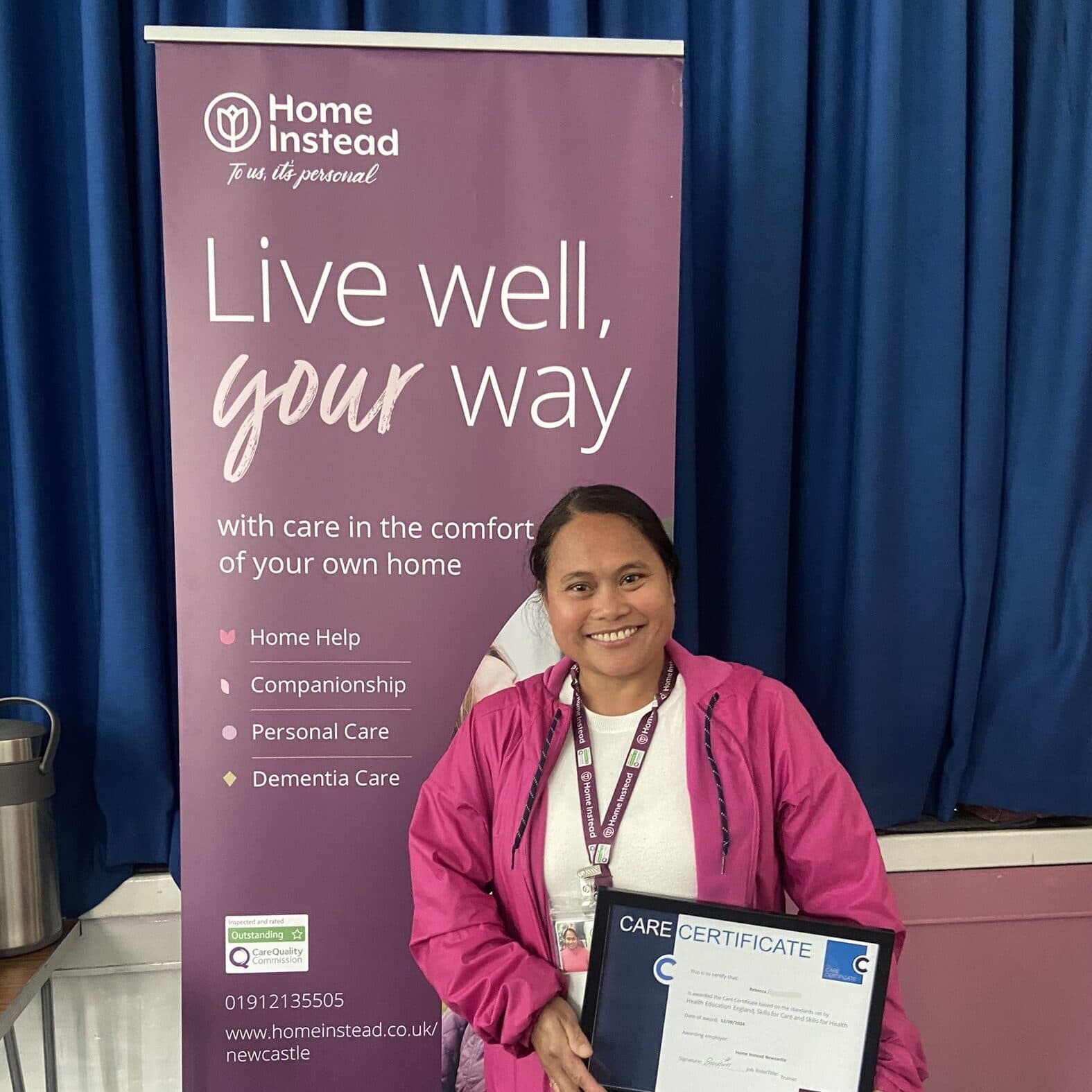 A smiling person in a pink jacket holds a care certificate in front of a "Home Instead" banner. - Home Instead
