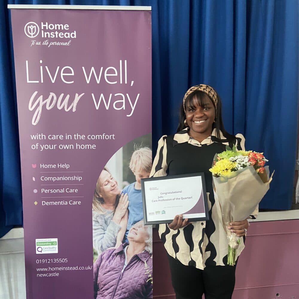 Person smiling, holding a certificate and flowers, standing next to a "Home Instead" banner. - Home Instead