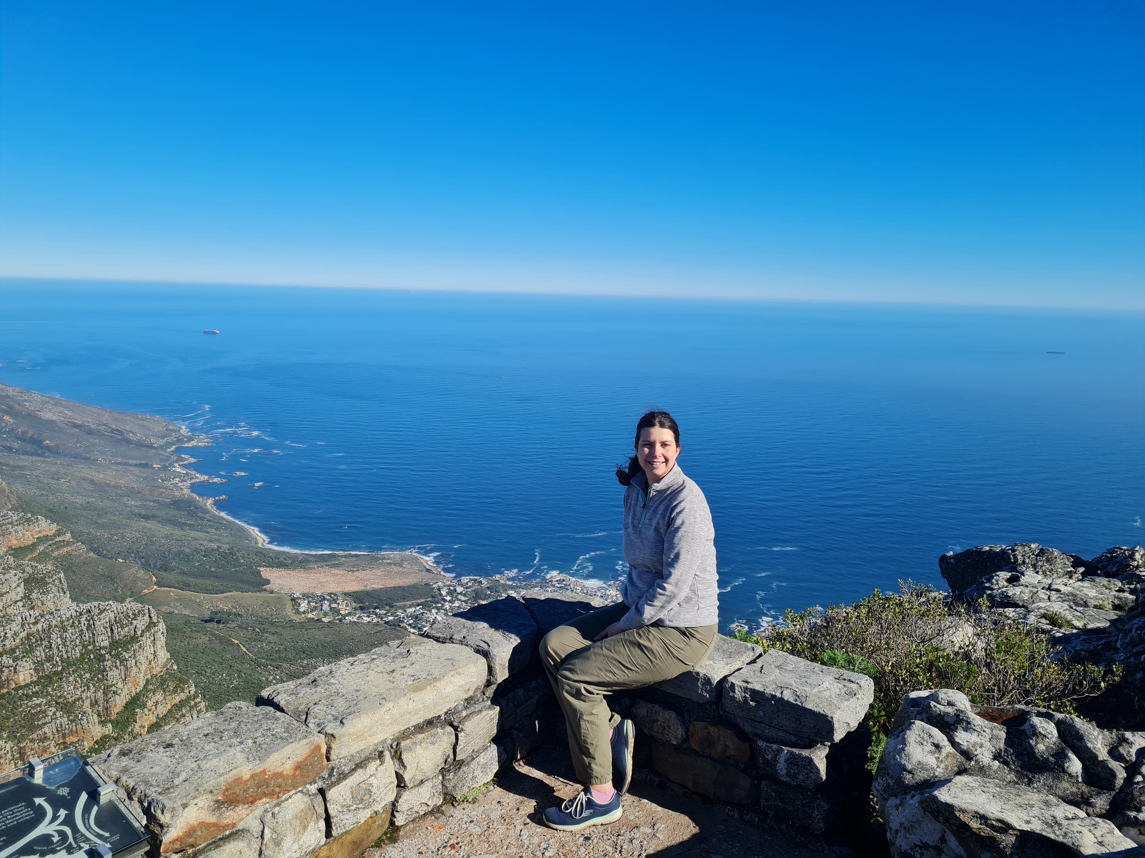A person sitting on a stone ledge overlooking a vast ocean and coastal landscape under a clear blue sky. - Home Instead