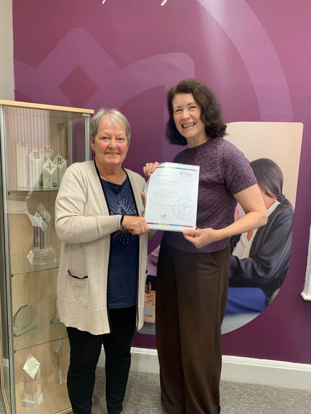 Two women smiling and holding a certificate in front of a purple wall and display case with awards. - Home Instead