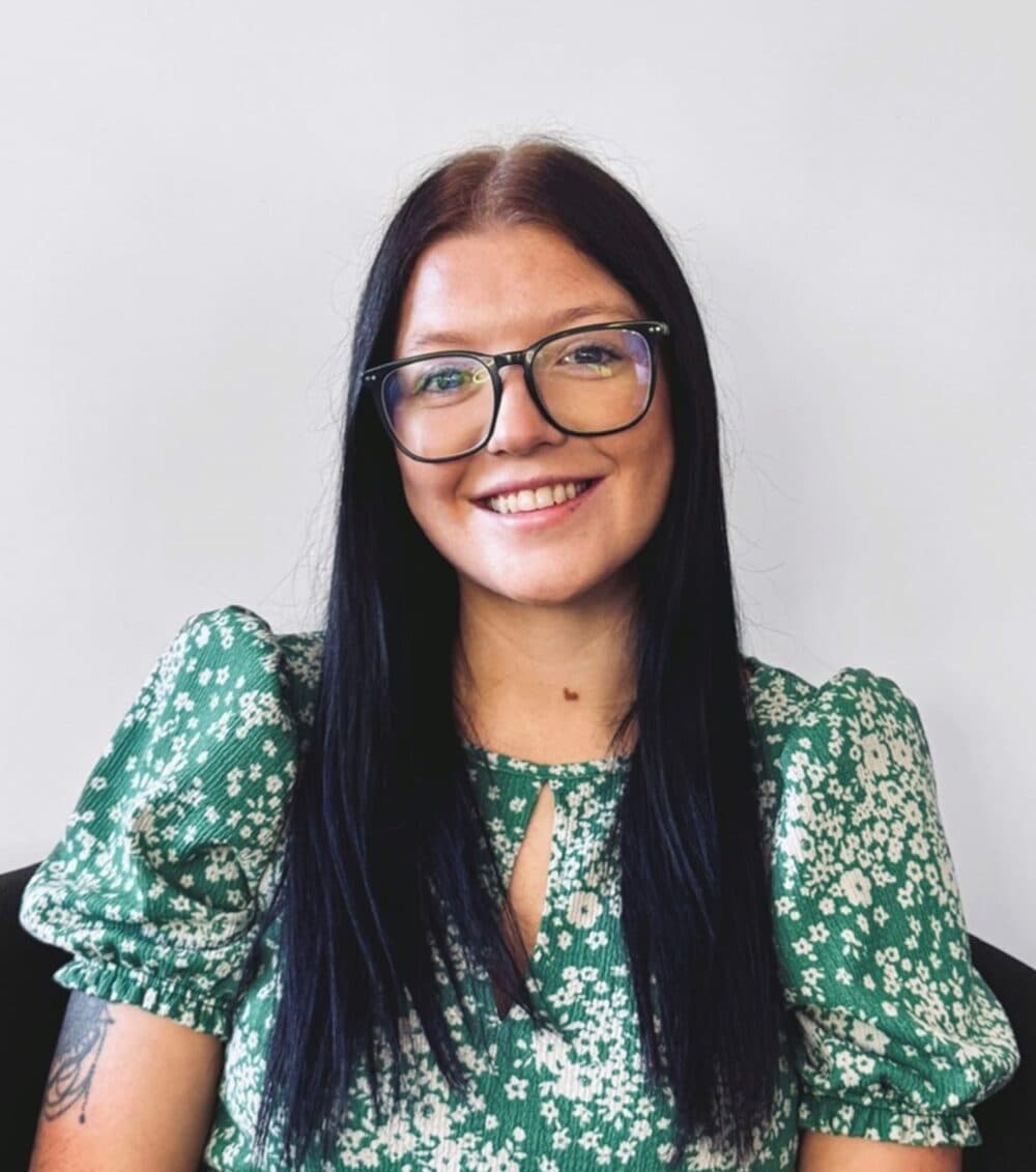 Woman with long dark hair and glasses, smiling, wearing a green floral dress, sitting against a plain background. - Home Instead