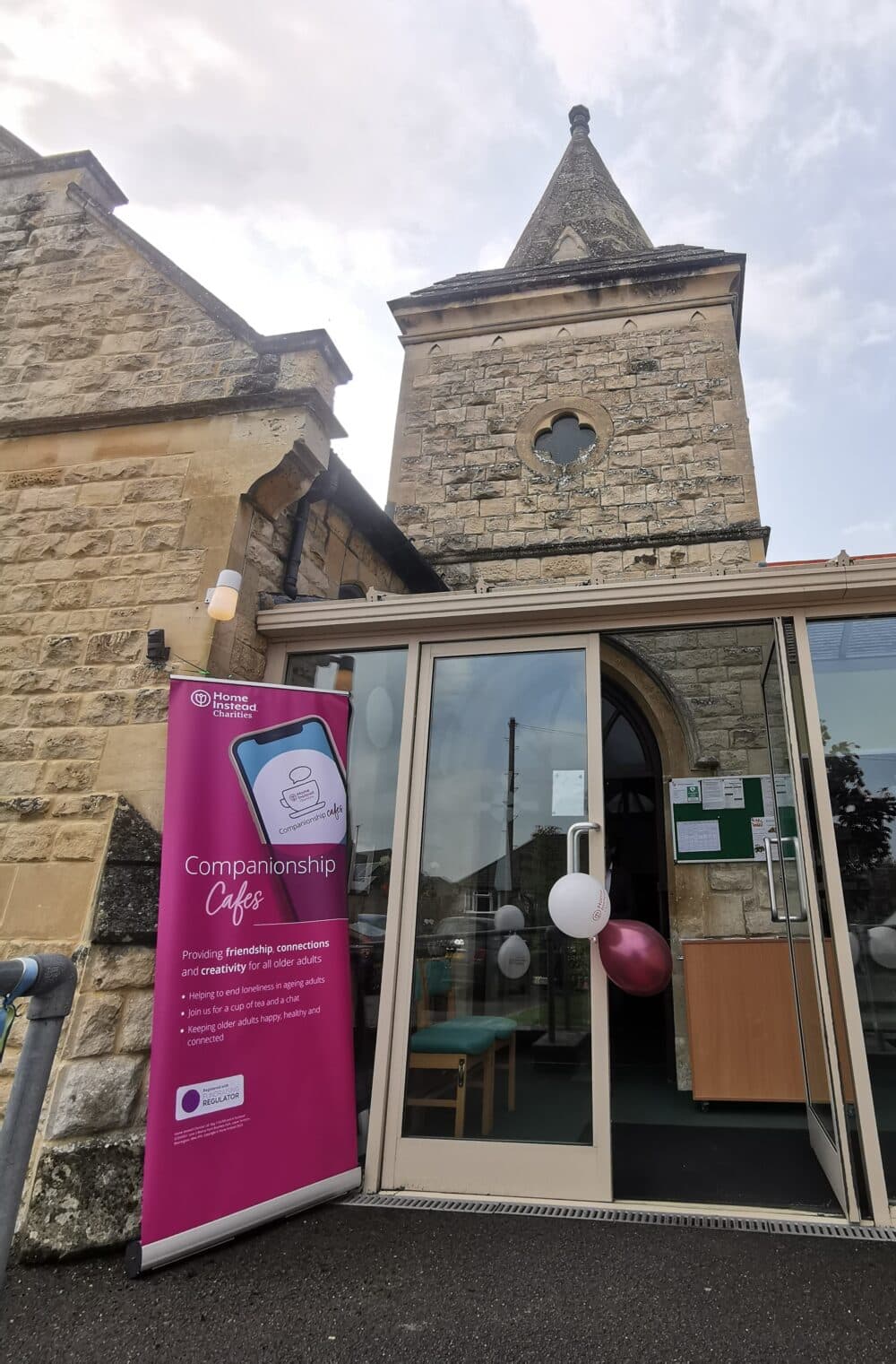Entrance to Churchdown Methodist Church with a Companionship Cafe banner outside and balloons on the door handle. The banner reads "Home Instead Charities Companionship Cafes. Providing friendship, connections and creativity for all older adults".