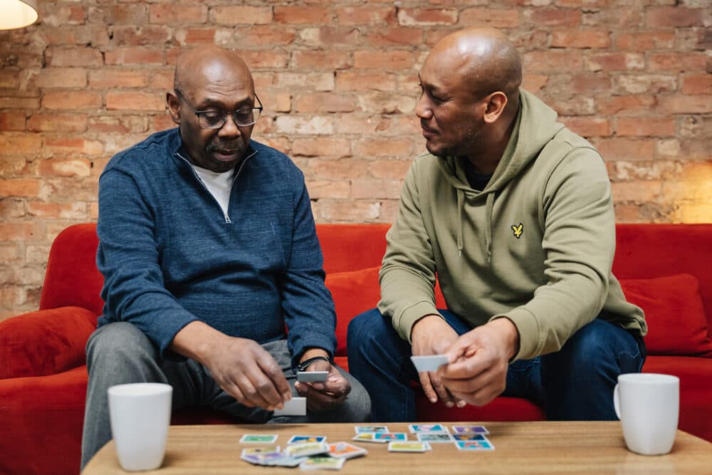 Two men sitting on a red couch, playing card games on a wooden table, with two white mugs beside them. - Home Instead