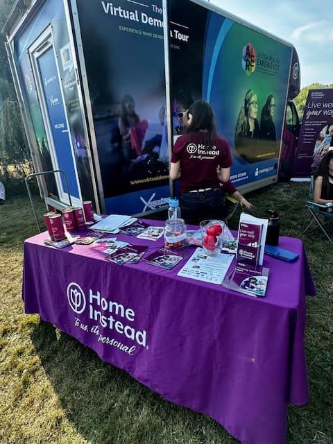 Booth at an event with Home Instead branding, displaying brochures and giveaway items on a table outdoors. - Home Instead