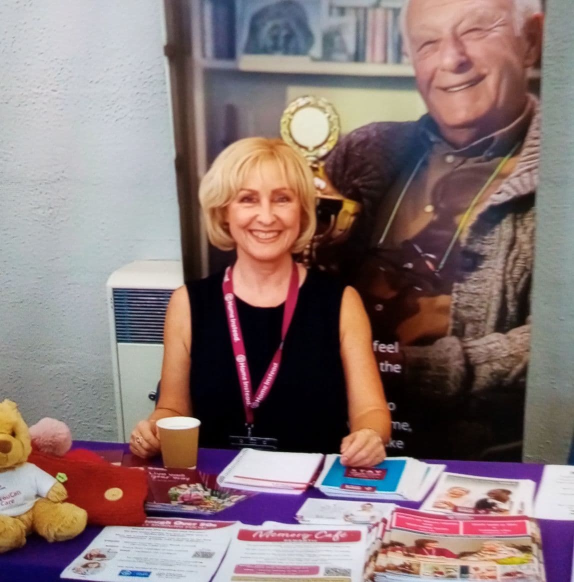 Woman smiling at an information booth with pamphlets and a teddy bear on a purple tablecloth. - Home Instead