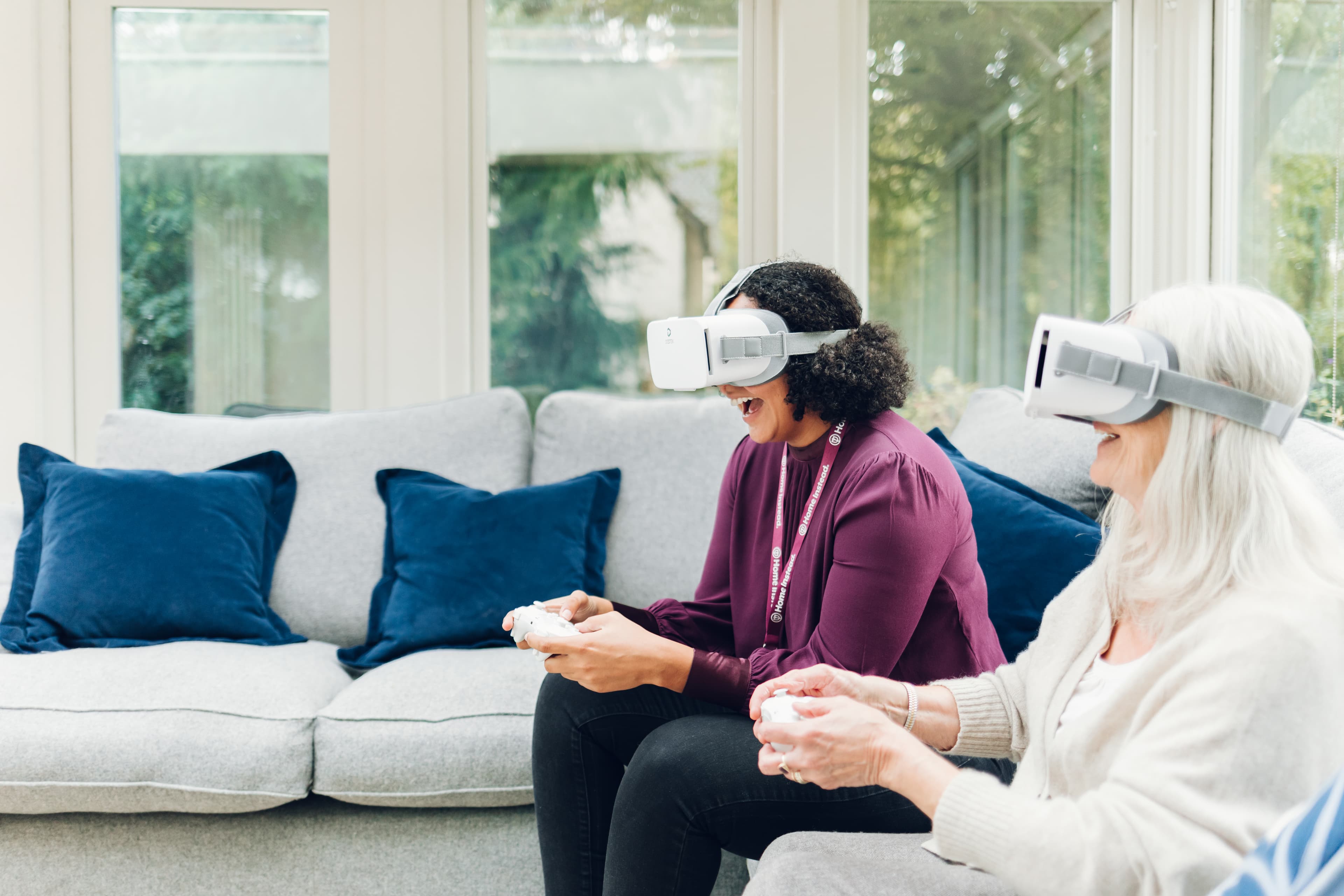 Two people sitting on a sofa, wearing VR headsets and holding controllers, engaged in a virtual reality experience. - Home Instead